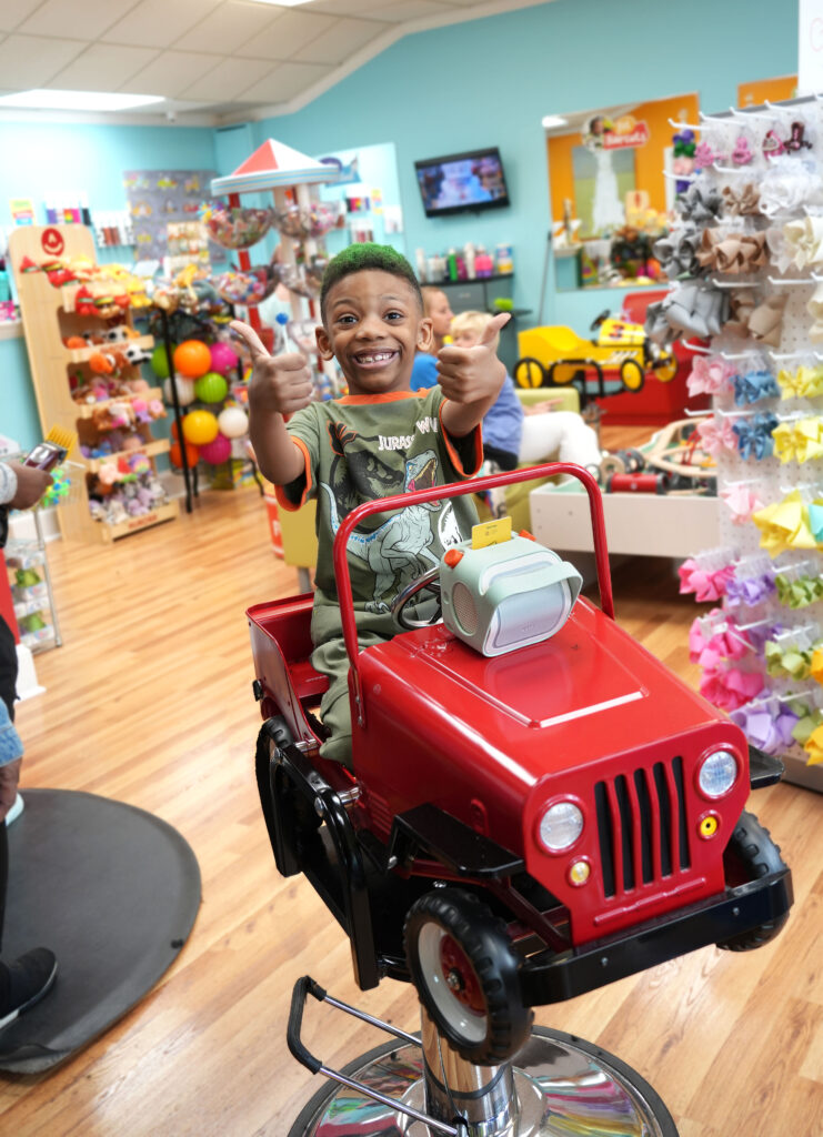 A happy young boy flashes a big smile and gives two enthusiastic thumbs up while seated in one of our fun vehicle-themed styling chairs at Pigtails & Crewcuts Buckhead. He has a Yoto player with him, adding extra joy to his haircut experience!