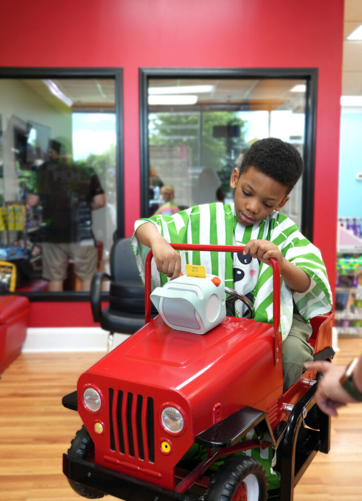 A young boy sits in a vehicle-themed styling chair at Pigtails & Crewcuts Buckhead, happily inserting a card into the Yoto player.