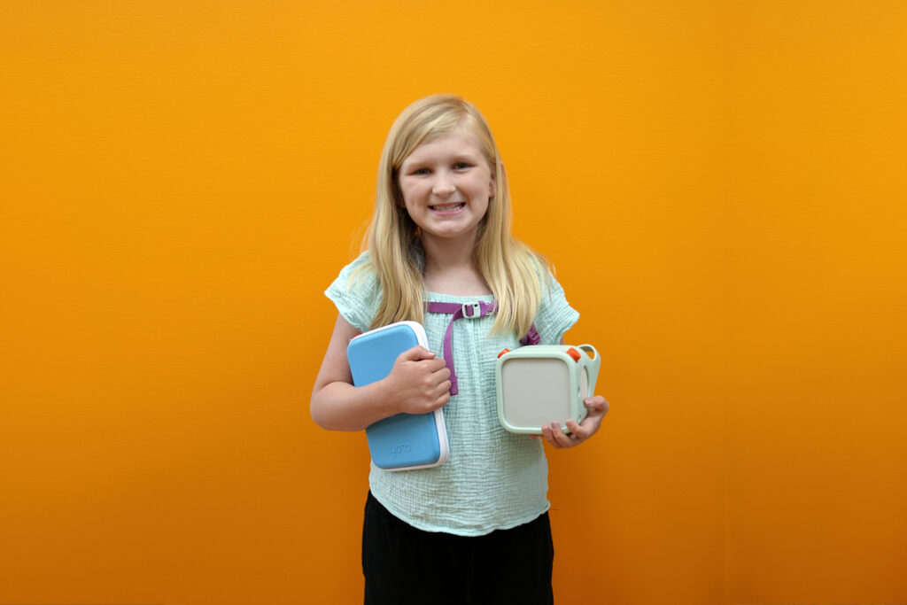 A smiling blonde girl holds a Yoto player and matching Yoto card case at Pigtails & Crewcuts Buckhead.