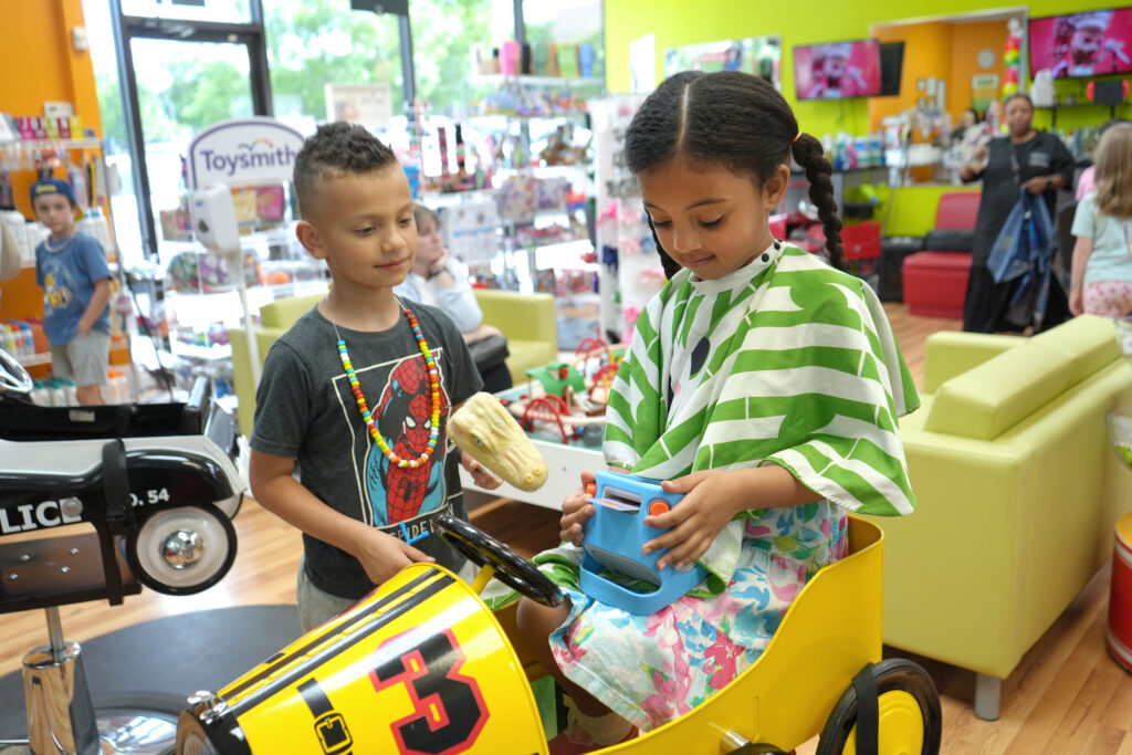 A girl sits in a vehicle-themed styling chair, playing with a Yoto player, as a young boy approaches to join her.