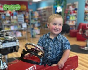 Smiling young child in a red fire truck salon chair at Pigtails & Crewcuts East Memphis after a back-to-school haircut.