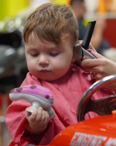 Toddler holding a toy while a stylist trims around the ear with clippers in a vehicle-themed salon chair.